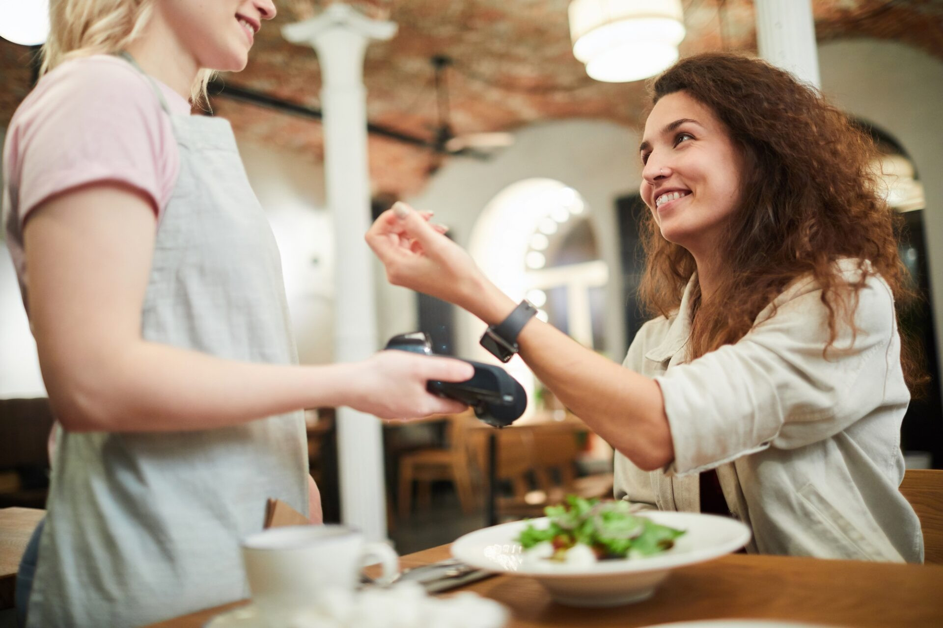 Joyeuse jeune femme excitée aux cheveux bouclés assis à table et payant pour dîner avec smartwatch tout en souriant à une serveuse amicale dans le restaurant