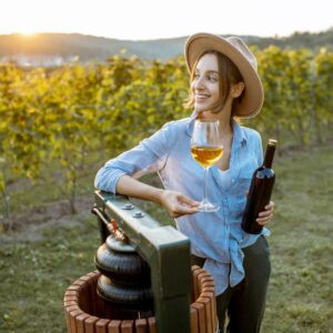 Femme jeune et joyeuse a goûté au vin, tout en se tenant près de la machine de presse sur le vignoble lors d'une soirée ensoleillée